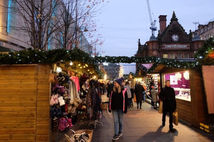 People browsing stalls at Glasgow Christmas Market in St Enochs Square with lights, decorations, and wooden huts selling clothes and gifts.