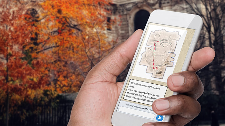 A hand holding a phone playing Treasure Hunt Glasgow with a tree with orange leaves behind.