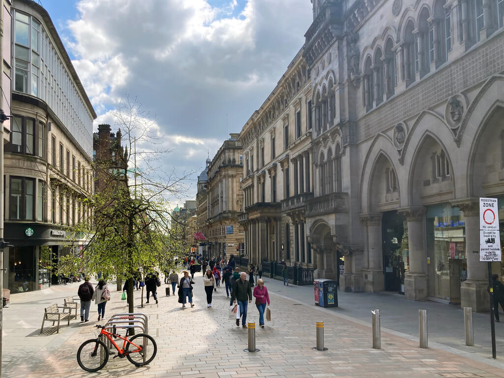 A bustling pedestrian street in Glasgow with diverse architecture, people walking, and an ornate stone building on the right
