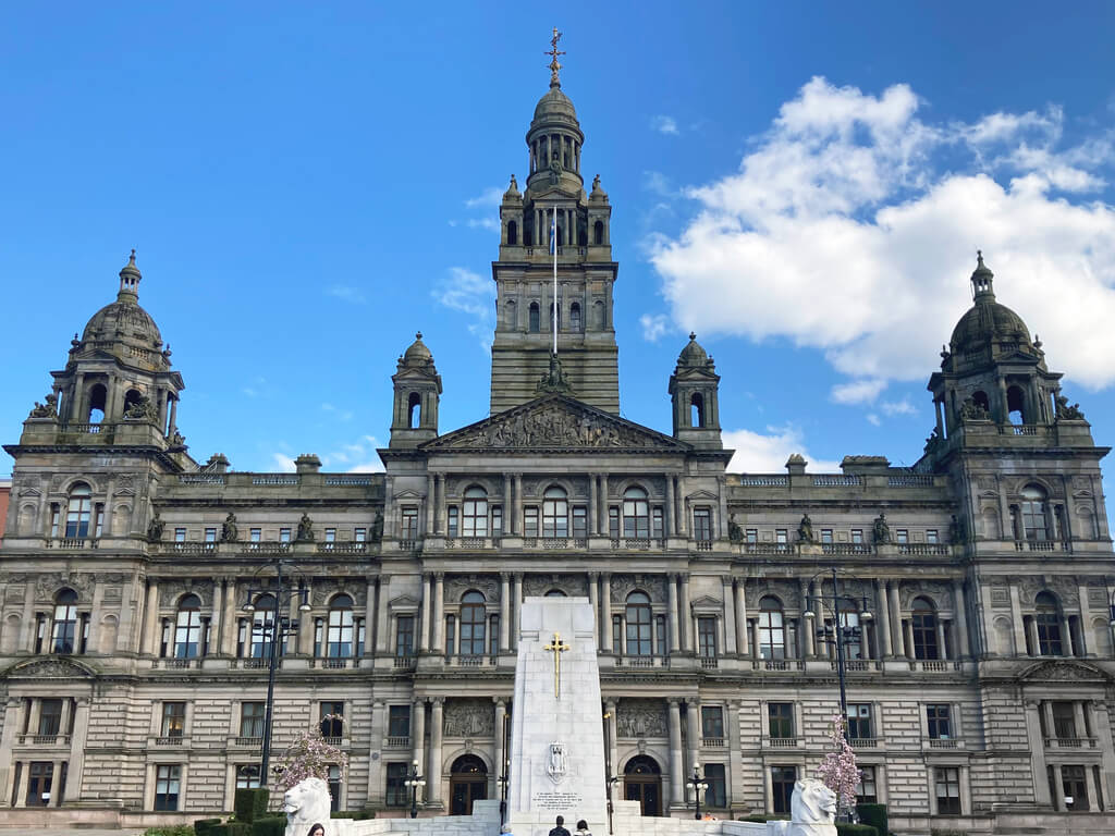 The imposing facade of Glasgow City Chambers with classical architecture and the Cenotaph war memorial in front, under a partly cloudy sky